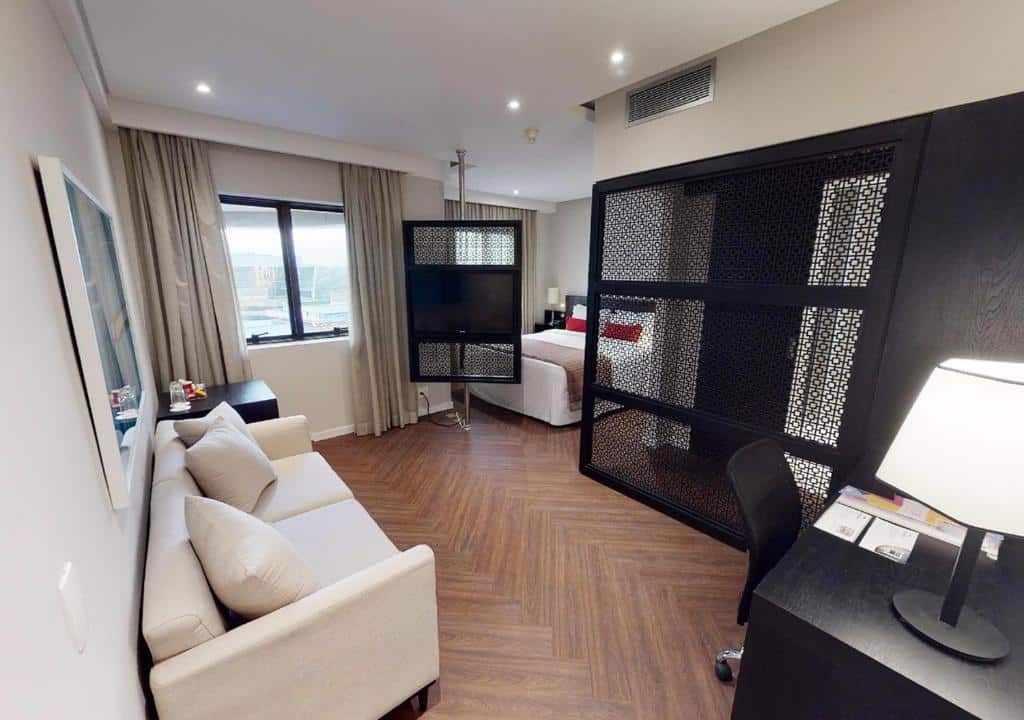 Vista do quarto do Estanplaza Funchal Faria Lima, na esquerda há um sofá bege claro com travesseiros e um quadro pendurado na parede. Na esquerda, uma escrivaninha com cadeira na cor preta, e um abajur está em cima da mesa. Um divisor preto separa a cama do restante do quarto. As duas janelas do quarto estão acompanhadas por cortinas beges. Há uma TV encaixada em uma plataforma giratória que pode ser virada de frente para a cama. Foto para ilustrar o post de hotéis na Faria Lima em SP.