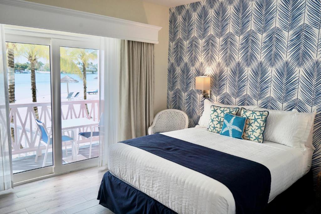 Quarto do Sandyport Beach Resort que mostra uma cama de casal com travesseiros, almofadas e cobertas. Do lado direito da cama tem uma luminária na parede e uma cadeira. Ao lado tem uma porta de vidro com cortinas que dá acesso a varanda do quarto com duas cadeiras e uma mesa com vista para o mar.