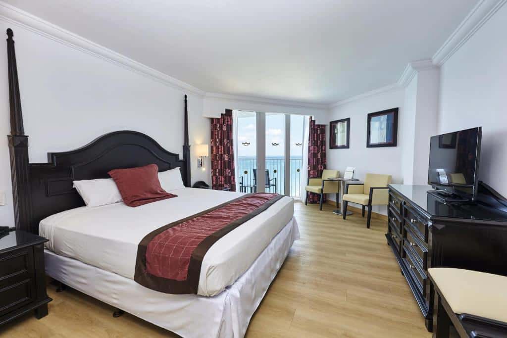 Quarto do Riu Palace Paradise Island com uma cama de casal ao centro do quarto com travesseiros e cobertas em cima. Do lado direito da cama tem uma mesa de cabeceira e em frente um cômoda de madeira com uma televisão em cima. Já do lado esquerdo do quarto tem duas poltronas e uma mesa, e mais ao fundo tem uma porta de vidro com cortinas que dá acesso a varanda do quarto.