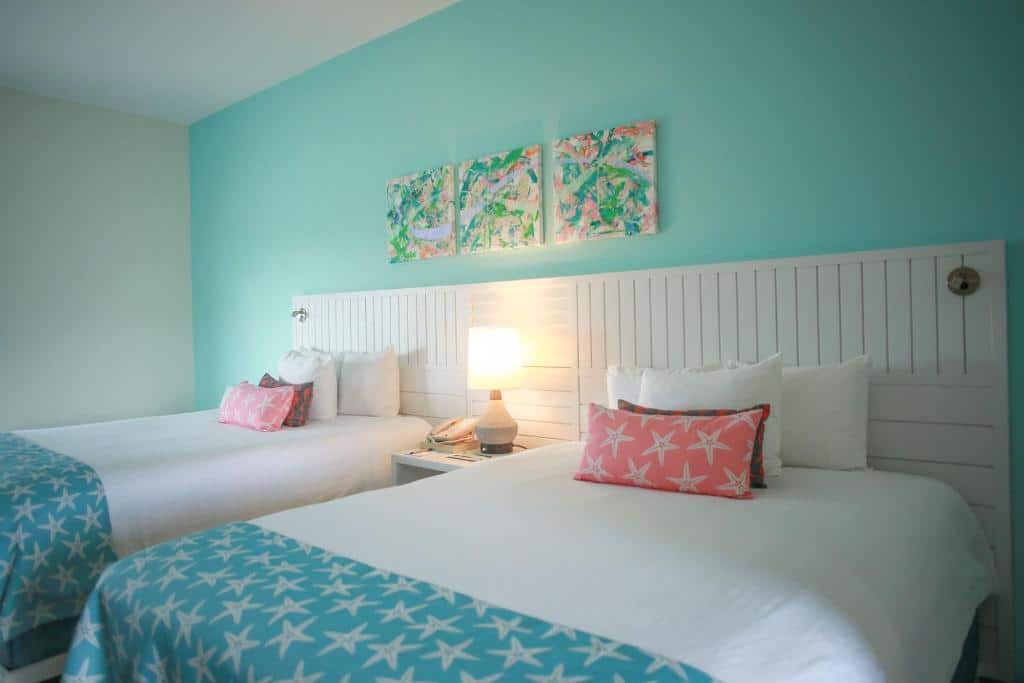 Quarto do Pelican Bay Hotel que mostra duas camas de solteiro lado a lado separadas apenas por uma mesa de cabeceira com um abajur em cima. As camas tem travesseiros, almofadas e cobertas, e atrás delas sua cabeceira é de madeira branca e tem três quadros na parede.