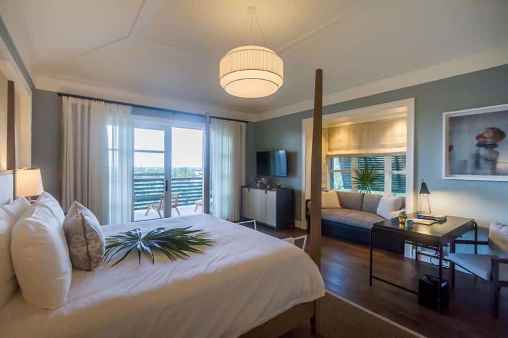 Quarto do The Island House com uma cama de casal do lado esquerdo do quarto com travesseiros, coberta e uma planta em cima. Já do lado direito do quarto tem uma parte que mostra uma mesa quadrada com uma cadeira, um sofá perto de uma janela e um cômoda com uma tv. Já do lado esquerdo da cama tem uma porta de vidro com cortinas que dá acesso a varanda do quarto. 