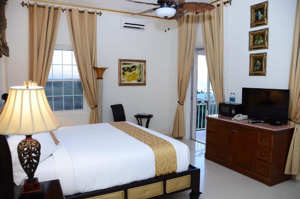 Quarto do Ocean West Boutique Hotel com uma cama de casal e do lado da cama uma mesa de cabeceira com um abajur. Em frente a cama tem uma cômoda com alguns objetos e uma tv em cima. No quarto também tem uma janela com cortinas, uma mesinha com uma cadeira e do lado da cômoda uma porta de vidro com cortinas que dá acesso a varanda do quarto.