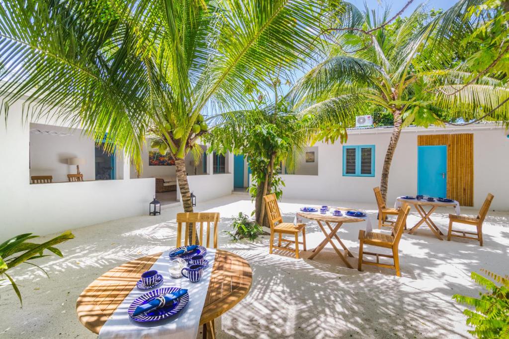 Área externa da Villa Rosa Maldives, um dos hotéis nas Maldivas, com mesas redondas e cadeiras espalhadas com xícaras e pratos azuis em cima. Há várias árvores deixando o espaço com sombra e clima aconchegante