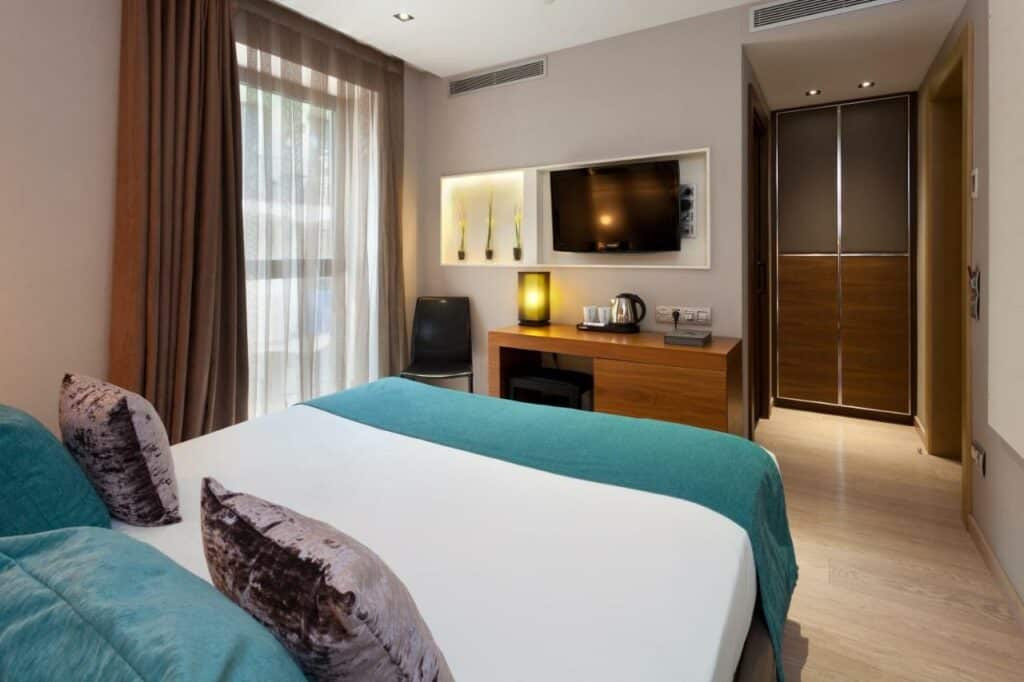 Quarto do Hotel España Ramblas. Uma cama de casal encara a televisão na parede acima de uma mesa de trabalho que tem banco, luminária e amenidades de chá. Há uma cadeira perto da janela que está coberta por cortinas marrom e brancas diáfanas. 