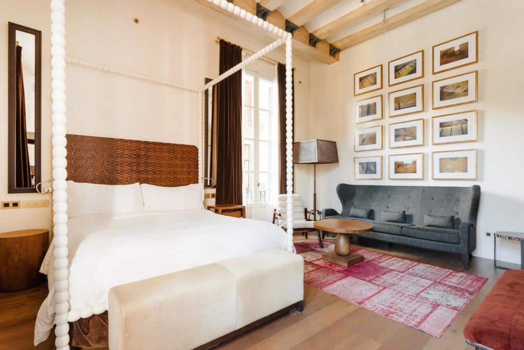 Quarto do Sonder DO Plaça Reial. Uma cama de casal com dossel branco tem um banquinho aos seus pés e mesinhas de cabeceira com tomada dos dois lados. Ao lado da cama há uma área de estar com sofá, mesa de centro, tapete e abajur. Diversos quadros pequenos estão dispostos na parede acima do sofá. Uma janela com cortinas marrom está atrás da poltrona e perto do abajur.