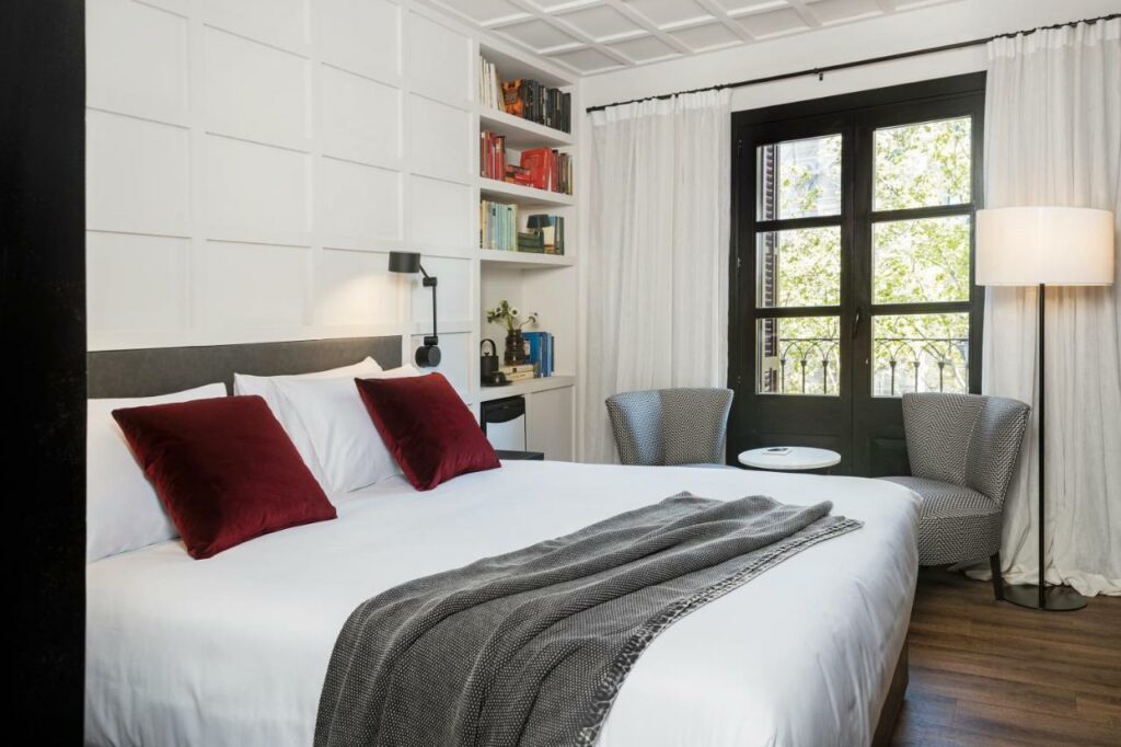 Quarto do Yurbban Ramblas Boutique Hotel. A cama de casal tem abajures dos dois lados, e uma estante com livros está ao lado direito da cama. Duas poltronas cinza com mesinha e um abajur estão em frente a uma janela com cortinas brancas em frente à estante.