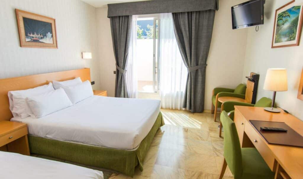 Quarto do Flor Parks, uma das recomendações de hotéis nas Ramblas em Barcelona. A cama de casal tem mesinhas de cabeceira dos ois lados e um quadro na parede acima. Há uma mesa de trabalho com cadeira, abajur e um controle remoto na parede em frente, e duas poltronas encaram a cama com uma mesinha entre elas. Há uma televisão encarando a cama. Ao fundo a janela tem cortinas cinza e brancas abertas.