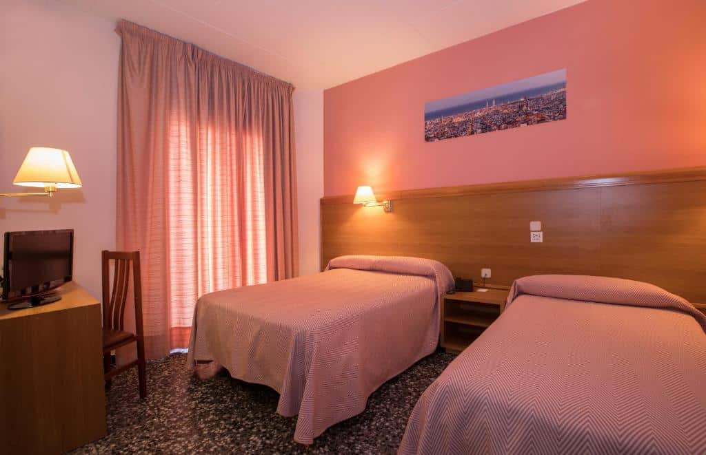 Quarto do Cuatro Naciones, uma das recomendações de hotéis nas Ramblas em Barcelona. Duas camas de solteiro estão separadas por uma mesinha de cabeceira e tem luminárias acima. Em frente, uma mesa de trabalho com cadeira e abajur tem também uma televisão que encara as camas. A janela atrás da mesa é coberta por cortinas bege.