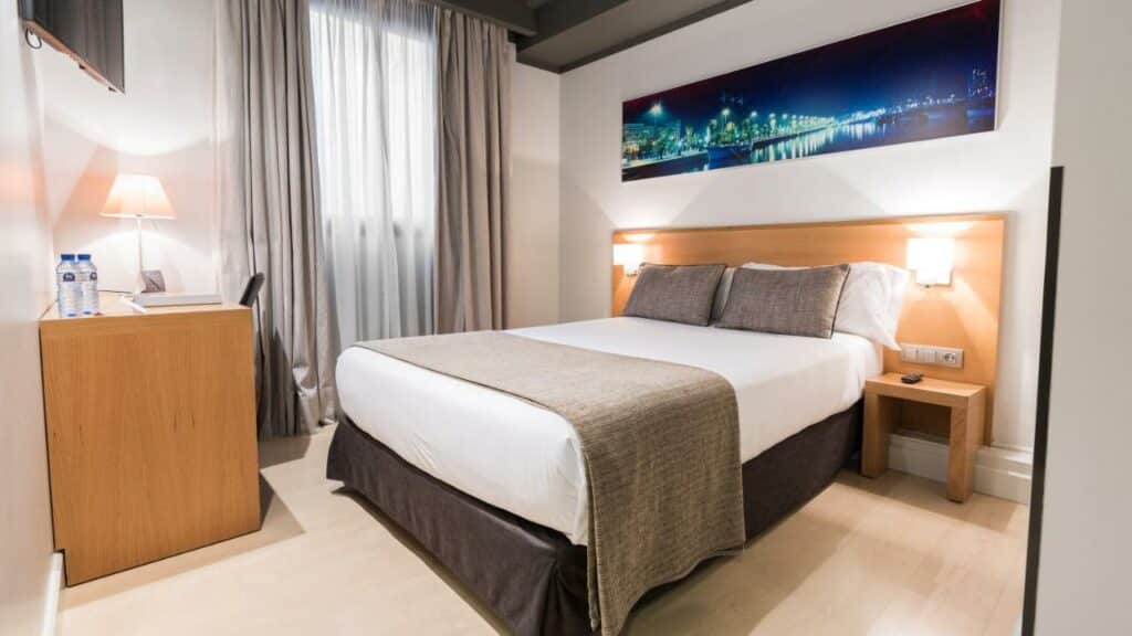 Quarto do Arc la Rambla, uma das recomendações de hotéis nas Ramblas em Barcelona. A cama de casal tem mesinhas de cabeceira dos dois lados com luminária fixada na cabeceira de madeira, além d eum quadro acima da cabeceira. Há uma televisão virada para a cama, uma mesa de trabalho ao lado tem abajur, cadeira e garrafas d'água. A janela está atrás da mesa e fica coberta com cortinas bege diáfanas.