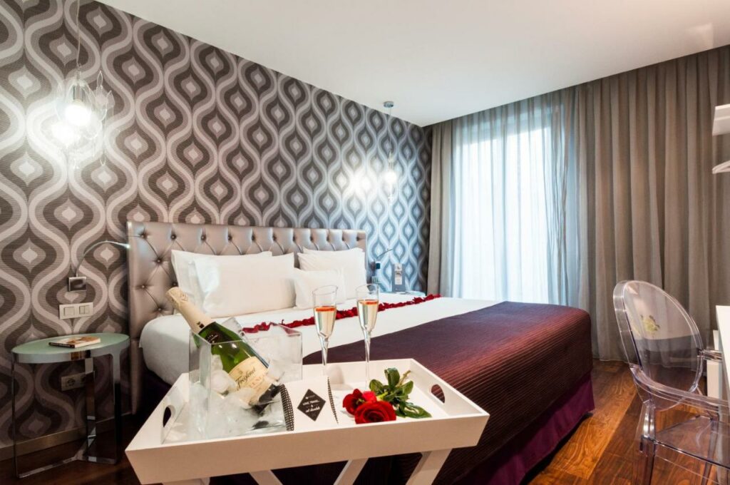 Quarto do Ikonik Ramblas, uma das recomendações de hotéis nas Ramblas em Barcelona. Com decoração romântica, o local tem uma cama de casal com rosas ao fundo e uma bandeja com garrafa de champanhe em um balde e um pouco servido em duas taças com rosas ao lado. Há mesinhas de cabeceira dos dois lados da cama, uma mesa de trabalho com cadeira em frente. Ao fundo, uma janela está coberta por cortinas bege diáfanas.