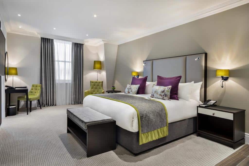 Quarto do St. James’ Court, A Taj Hotel, London com uma cama de casal, duas mesinhas de cabeceira, chão de madeira com um amplo tapete, há uma janela com cortinas e duas poltronas, para representar hotéis em Londres para brasileiros