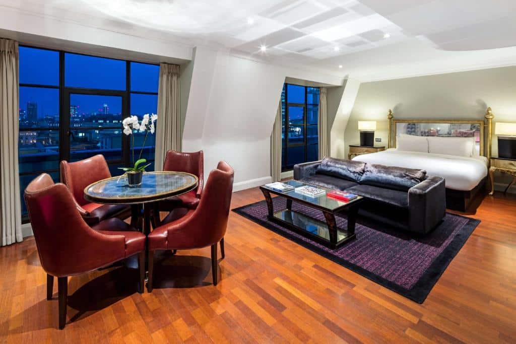 Quarto amplo do Radisson Blu Edwardian Hampshire Hotel, London com duas janelas panorâmicas, uma cama de casal com duas mesinhas de cabeceira, uma sofá com dois lugares e uma mesinha de centro, além de uma mesa redonda com quatro cadeiras, o piso é de madeira, para representar hotéis em Londres para brasileiros