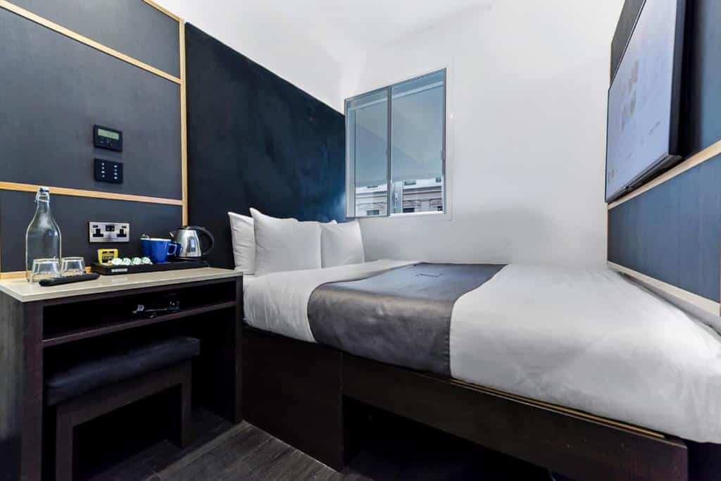 Quarto do The Z Hotel Trafalgar com uma cama de casal, uma pequena janela, uma televisão e uma pequena mesa com água, cafeteira e um banco
