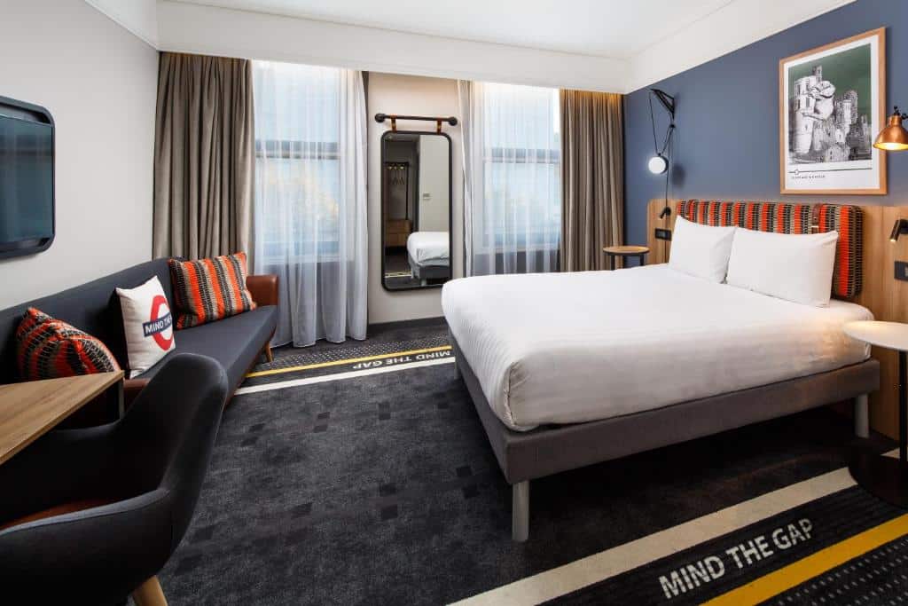 Quarto do ibis Styles London Gloucester Road com duas janelas com cortinas, um sofá com almofadas, uma mesa com uma cadeira, televisão e um espelho de corpo inteiro