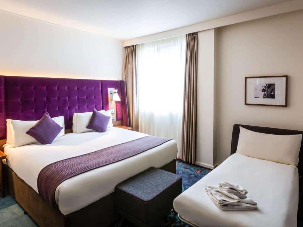 Quarto do Mercure London Bridge com uma cama de casal, uma de solteiro, uma janela com cortinas, um carpete colorido e uma cabeceira roxa