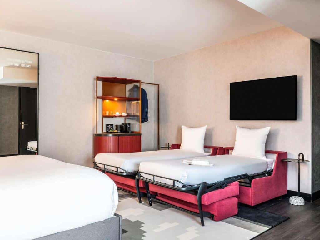 Quarto quadruplo do Tribe Amsterdam City, de 32 m², com cama de casal e a frente tem 2 sofás cama abertos com cochão e travesseitos acima. Há também uma TV e, ao lado, um móvel com amenidades para fazer chá ou café e um cabideiro. Representa hotéis para família em Amsterdam