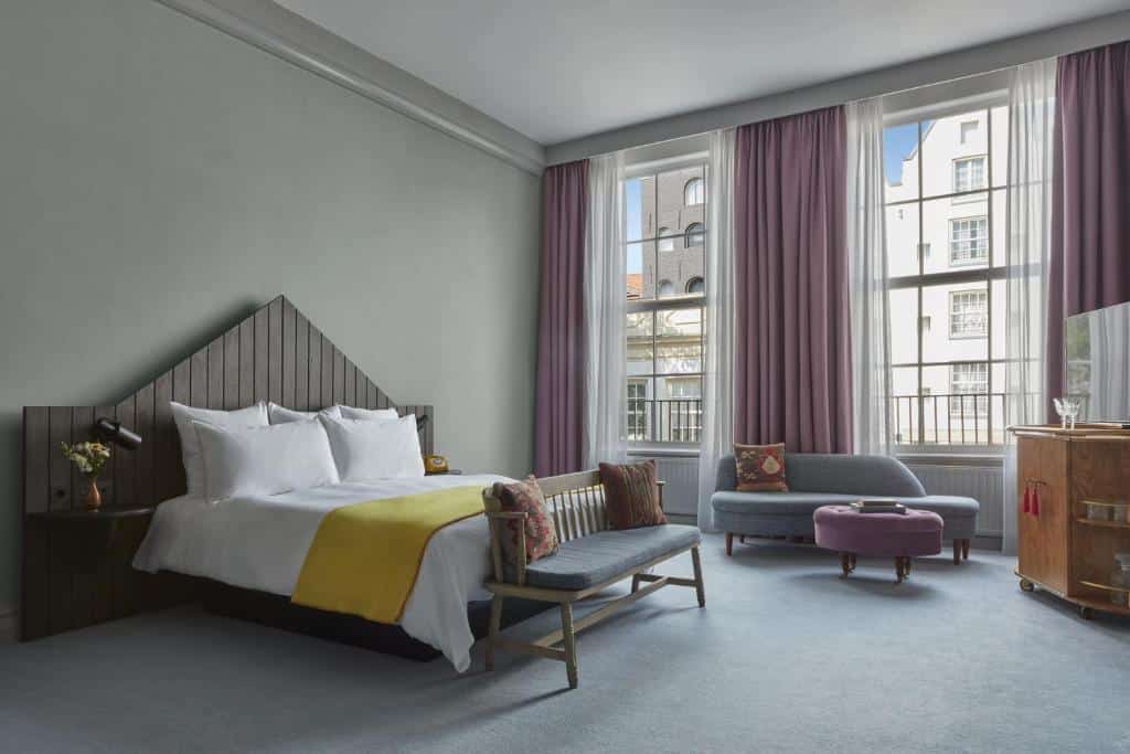 Quarto duplo do Pulitzer Amsterdam, de 30 m², com cama de casal, um banco a frente, e no lado direito tem um sofá, mesinha redonda estofada, e um móvel, além de janelas com cortinas lilás e branca. Representa hotéis para família em Amsterdam