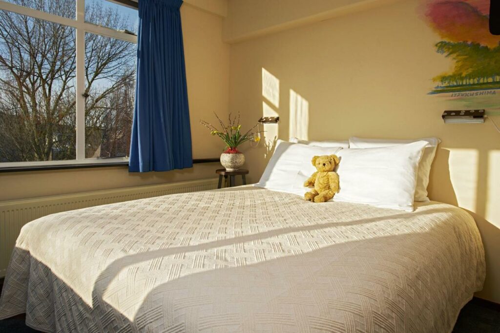 Cama de casal com um ursinho encostado nos travesseiros que estão em cima da cama, e no lado esquerdo tem uma mesinha redonda com um vaso de flor e uma janela ao lado com vista para árvores e uma cortina azul recolhida nos lados. Imagem para ilustrar o post de hotéis para família em Amsterdam