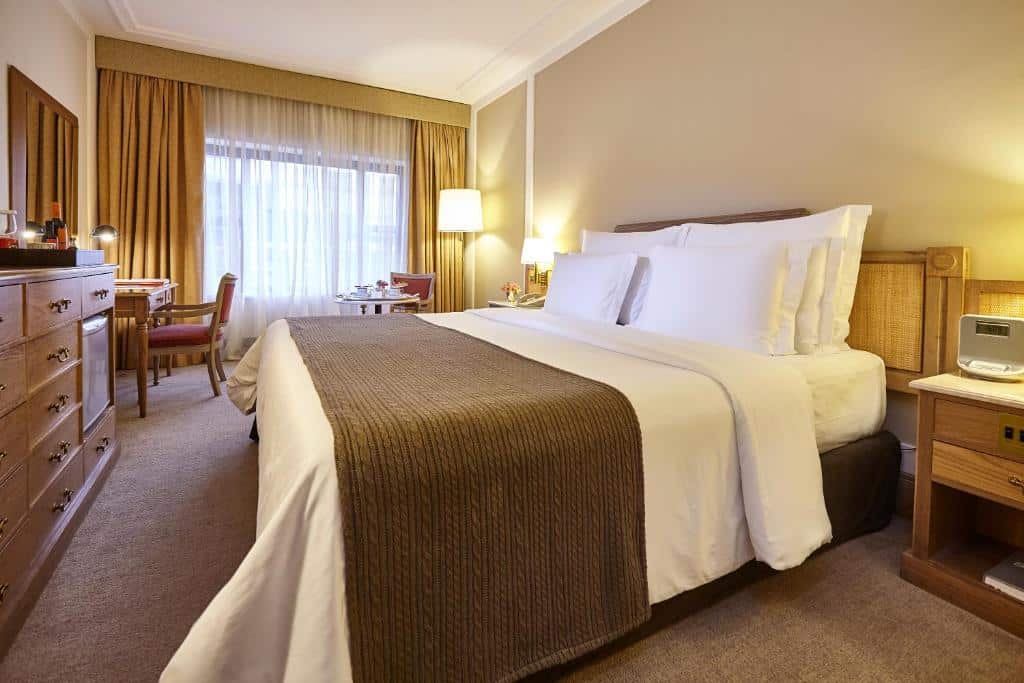 Quarto do L'Hotel PortoBay São Paulo, um dos hotéis perto da Avenida Paulista, com uma cama grande, cobertor marrom, cômoda, escrivaninha, cadeiras, luminárias e uma janela com cortinas.