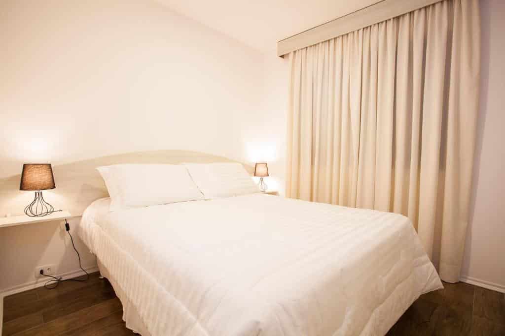 Uma cama bem arrumada com roupas de cama brancas, duas luminárias de cabeceira e cortinas bege cobrindo uma janela em um quarto do Travel Inn Park Avenue Jardins, um dos hotéis perto da Avenida Paulista.