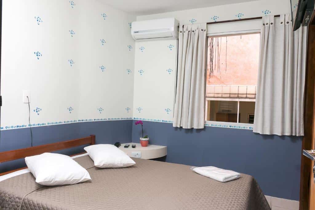 Pequeno quarto do Hotel Residenza, um dos hotéis perto da Avenida Paulista, com uma cama, dois travesseiros, toalha dobrada, mesa de cabeceira, janela com cortinas e ar-condicionado.