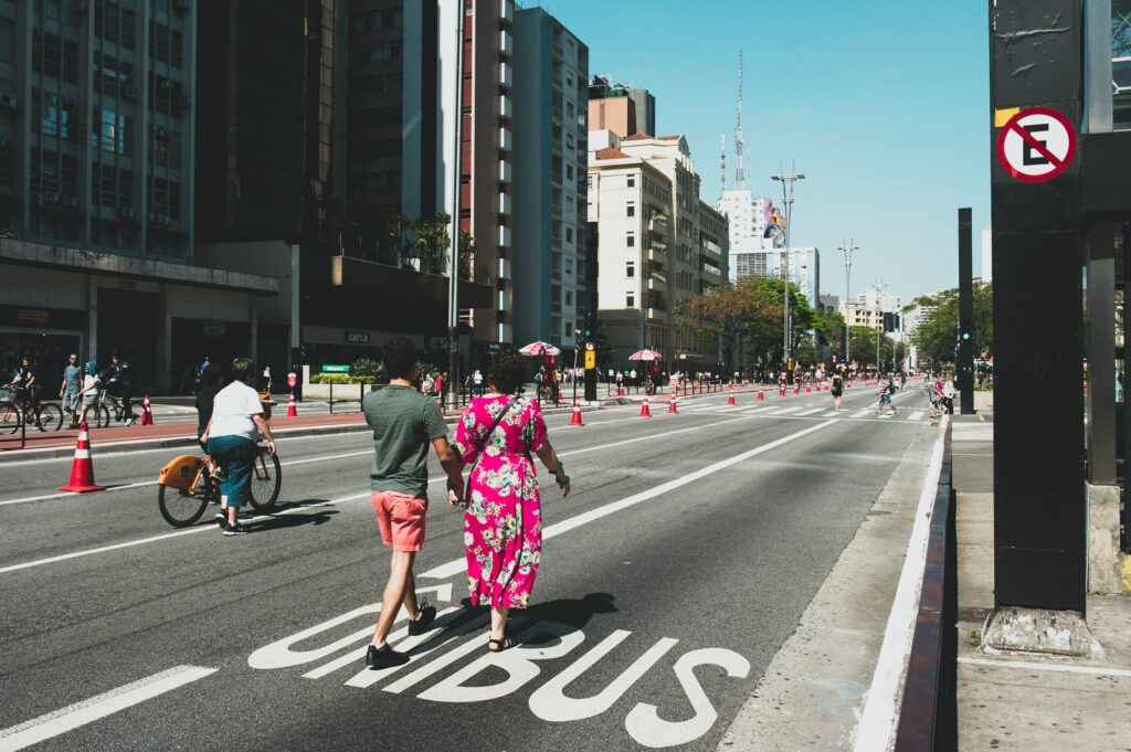 Um casal atravessa a Avenida Paulista, em São Paulo, em uma parte marcada para ônibus, com ciclistas e edifícios ao fundo em um dia ensolarado.