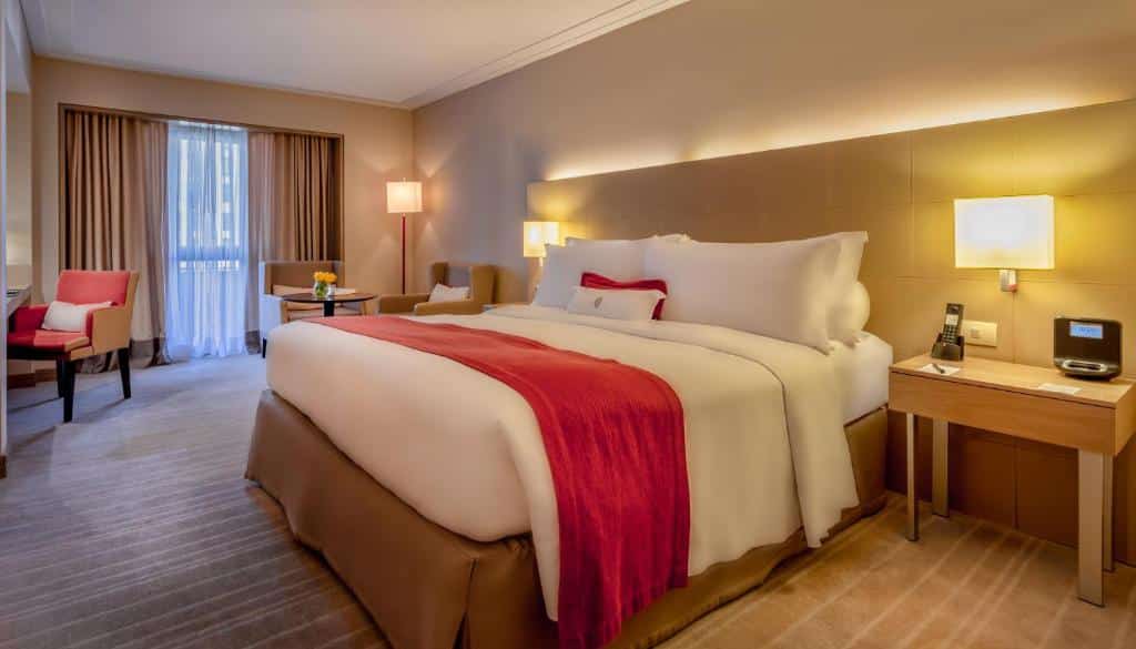 Quarto do InterContinental São Paulo, um dos hotéis perto da Avenida Paulista, com uma cama grande, corredor vermelho, mesinhas de cabeceira, cadeiras, escrivaninha e janela com cortinas que deixam entrar a luz do dia.