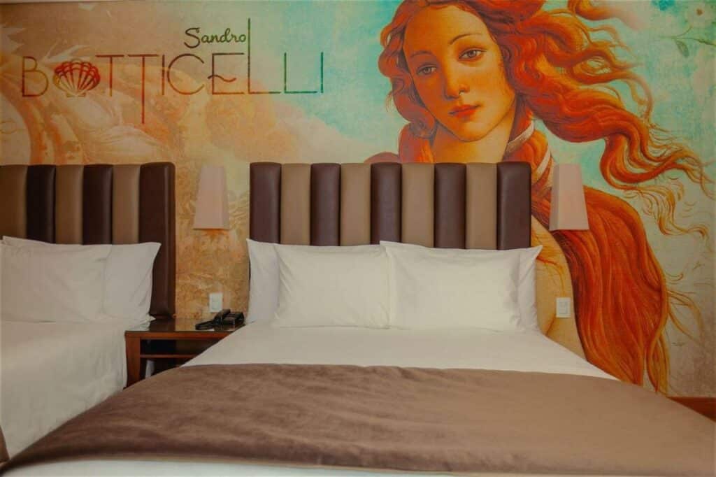Quarto do Vila Galé Paulista, com duas camas, lençóis brancos, cabeceiras acolchoadas e um mural da obra de arte de Sandro Botticelli acima das camas.