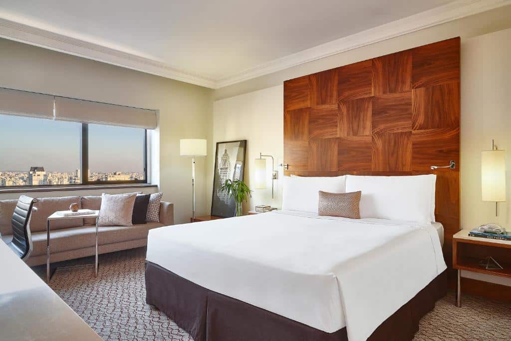 Quarto do Renaissance São Paulo Hotel, um dos hotéis perto da Avenida Paulista, sendo ele moderno com uma cama grande, sofá, escrivaninha e janela com vista para a cidade ao fundo.