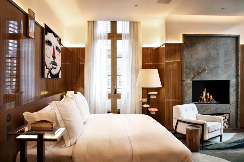 Quarto do Rosewood São Paulo com cama, poltrona, lareira, janela grande e uma obra de arte com retrato em uma parede com painéis de madeira.