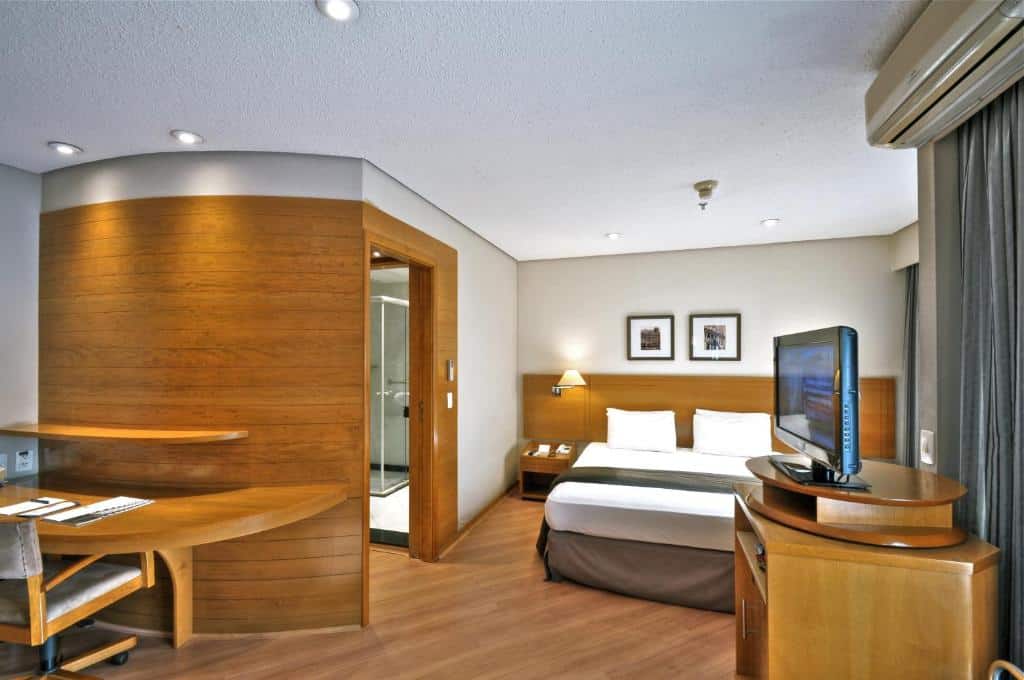 Quarto de um hotéis perto da Avenida Paulista, o Transamerica Executive Paulista, sendo ele moderno e com móveis de madeira, cama de casal, escrivaninha, TV e vista para um banheiro com chuveiro de vidro.