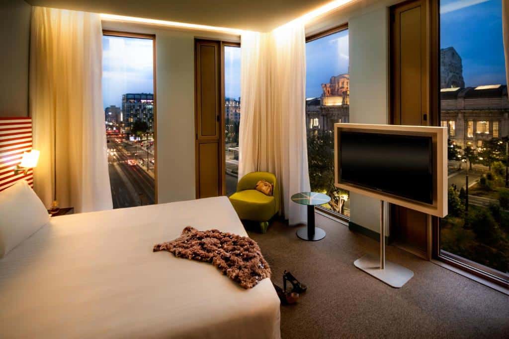 Quarto no Glam Milano com janelas panorâmicas, com uma televisão, uma cama de casal, uma poltrona e uma mesinha de centro, há cortinas brancas nas amplas janelas