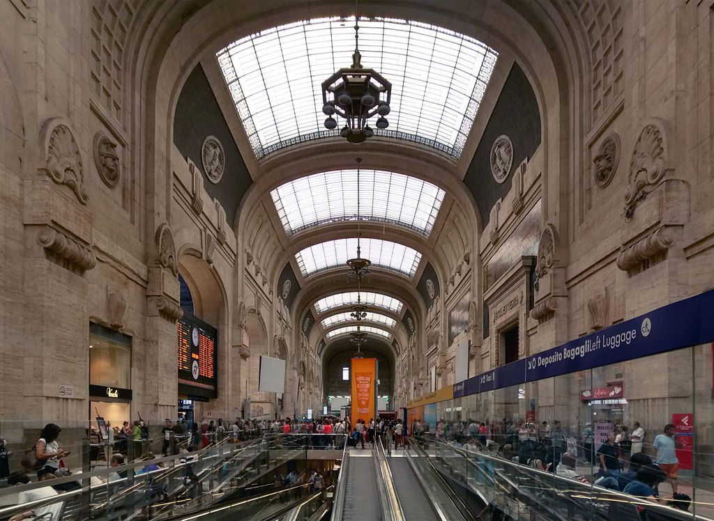 Lado interno da Estação Central, o prédio é muito alto com um teto de vidro, há diversas escadarias, muitas pessoas caminhando e a decoração das paredes lembra o renascentismo 