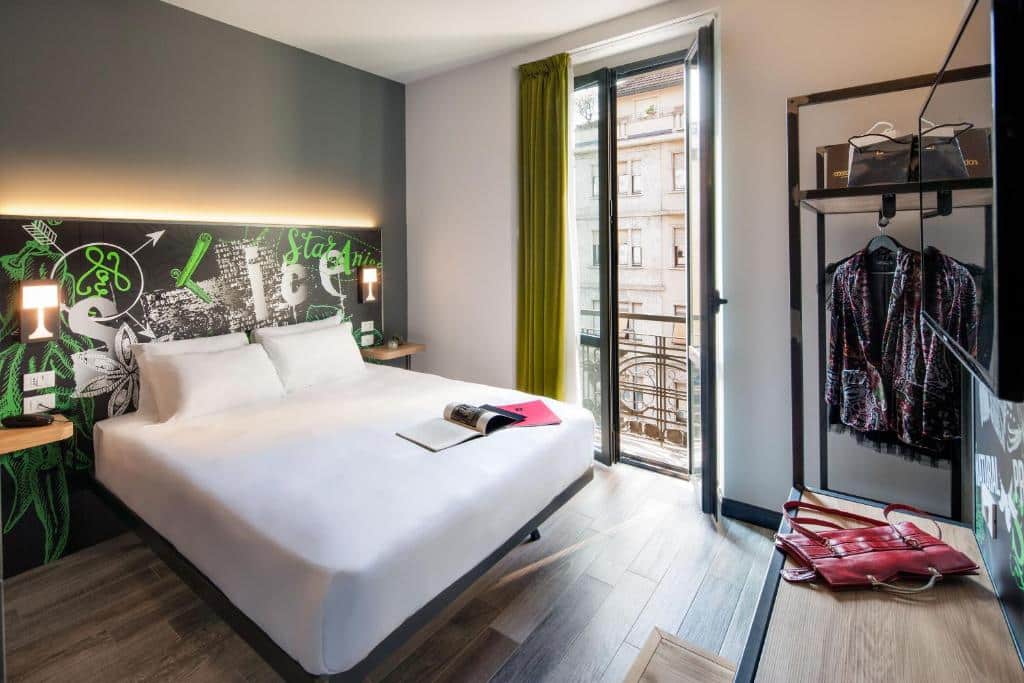 Quarto do Spice Hotel Milano com uma pequena varnada com portas de vidro e cortinas, uma cama de casal com uma cabeceira desenhada com luminárias presas nela, há um banco de madeira, uma televisão e um pequeno armário de conceito aberto com espaço para cabides