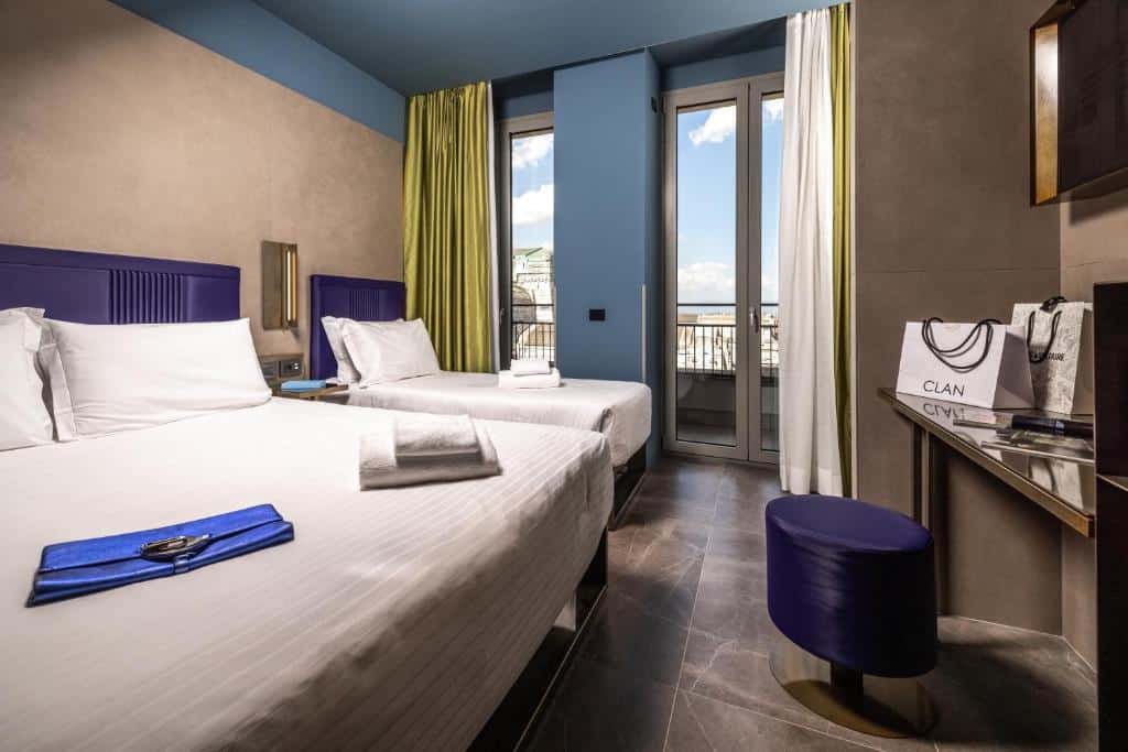 Quarto no HD8 Hotel Milano com portas de vidro com cortinas que levam para uma pequena varanda, há uma cama de casal e uma de solteiro, do lado direito, há uma pequena mesa de escritório e um bufe roxo, além de uma televisão na parede