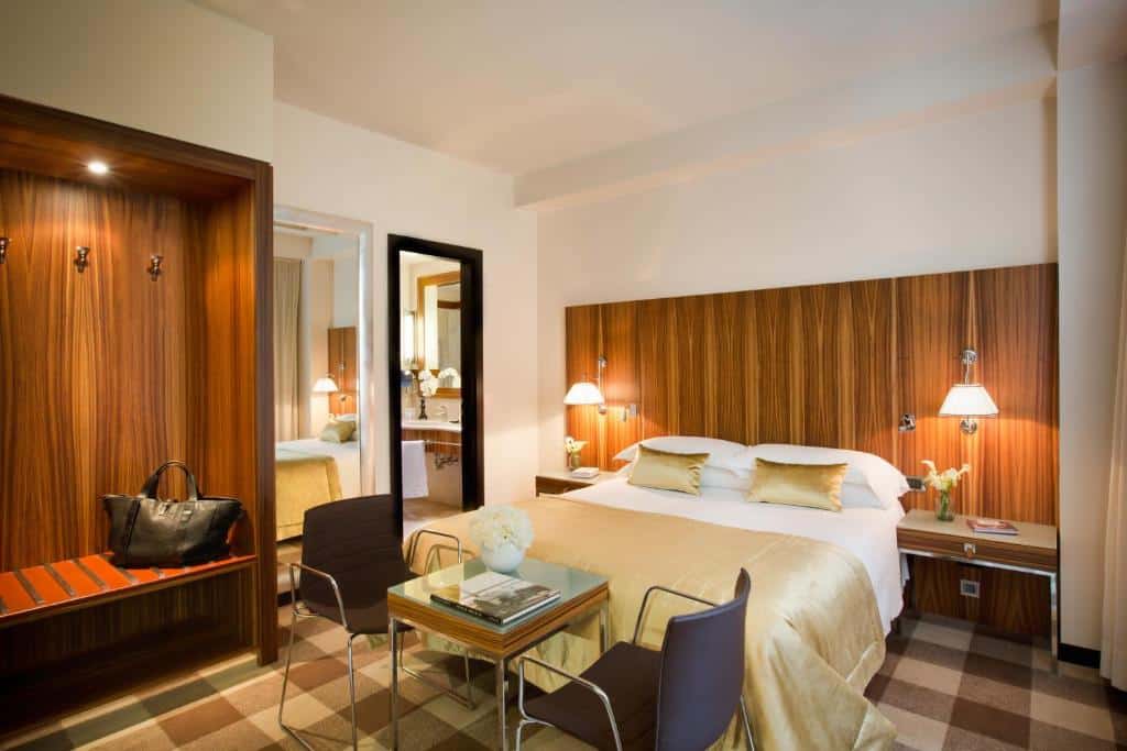 Quarto do Starhotels Anderson com uma cama de casal ampla, uma cabeceira de madeira com abajures presos, uma pequena mesa quadrada com duas cadeiras estão ao pé da cama, há também, do lado esquerdo, um armário sem portas e um espelho de corpo inteiro