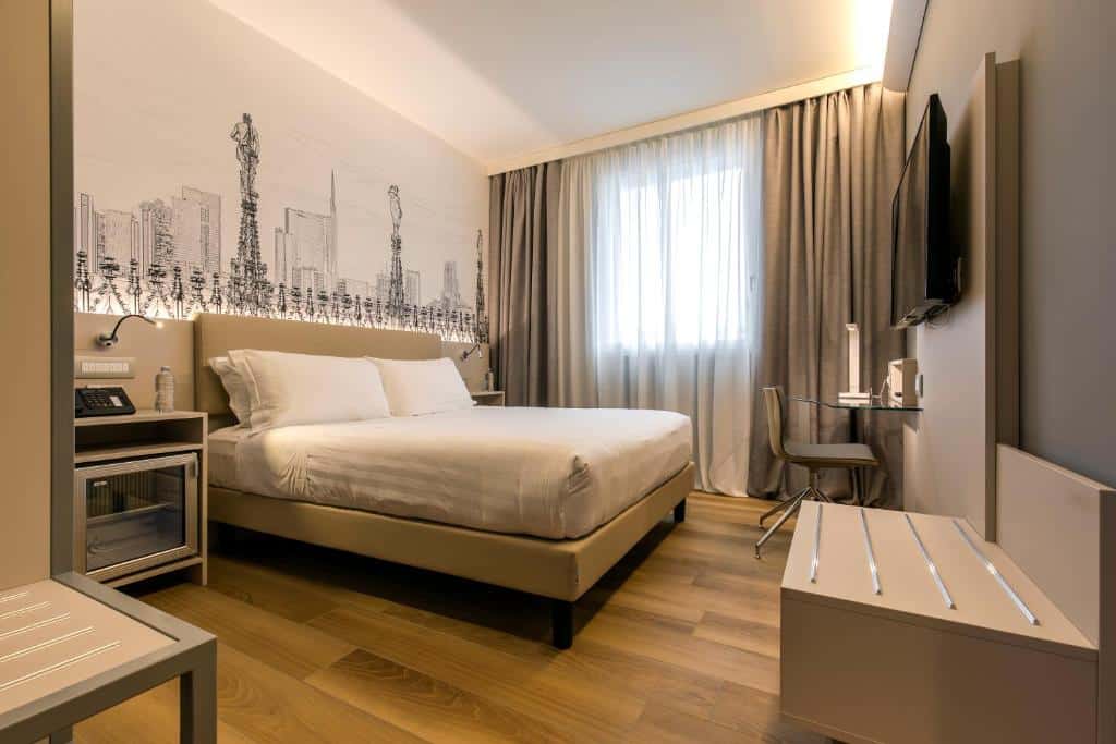 Quarto do 43 Station Hotel com uma cama de casal, uma cabeceira com papel de parede e luminárias presas, uma janela com cortinas, uma televisão, uma pequena mesa com uma cadeira giratória e um banco de madeira