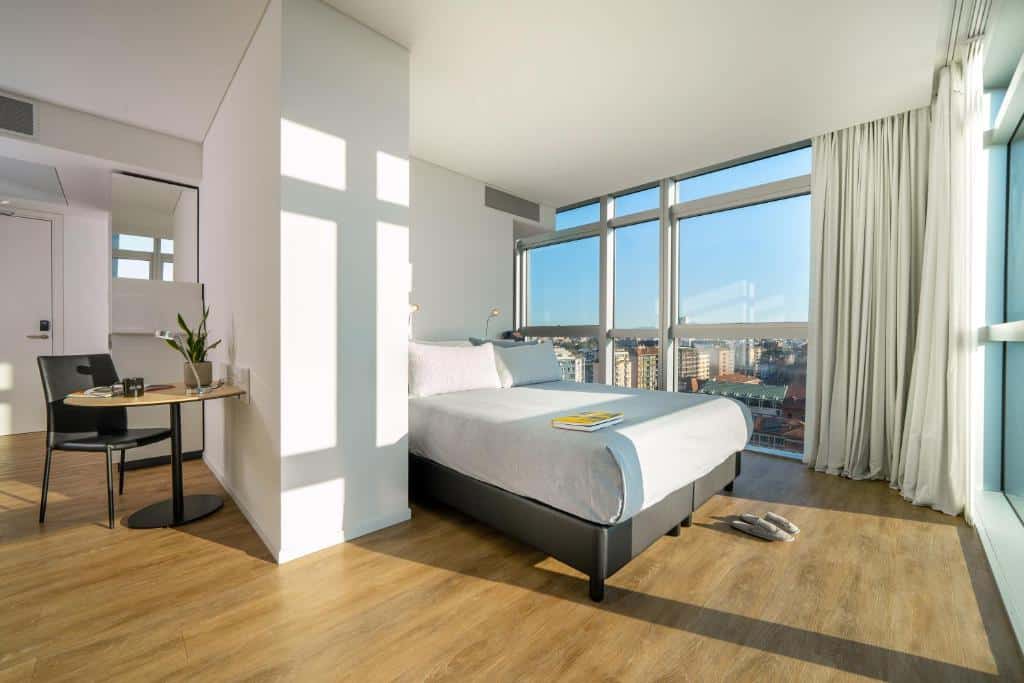 Quarto espaçoso do INNSiDE by Meliá Milano Torre GalFa com janelas panorâmicas com vista para a cidade, há uma cama de casal, o chão imita madeira, do lado esquerdo, é possível ver uma mesa redonda e uma cadeira