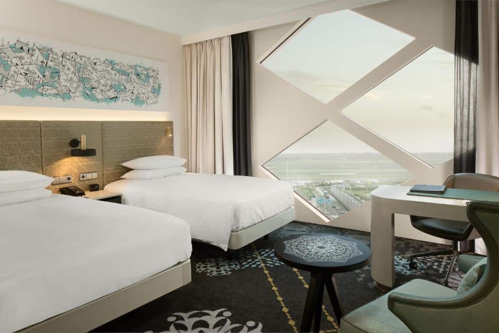 Quarto executivo com duas camas de solteiro do Hilton Amsterdam Airport Schiphol, de 29 m², com uma mesinha redonda, uma poltrona verde e uma mesa de trabalho com cadeira de escritório a frente. Ao lado há uma parede algumas partes de vidro no formato de triangulo com vista para a pista do aeroporto. Imagem para ilustrar o post de hotéis com vista do aeroporto em Amsterdam