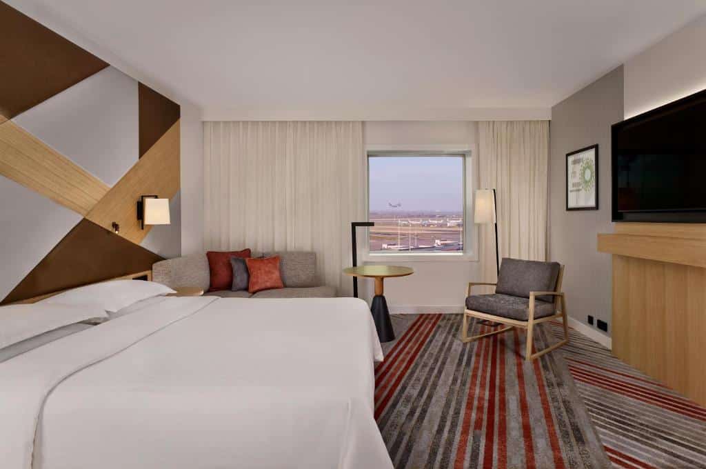 Quarto deluxe do Sheraton Amsterdam Airport Hotel and Conference Center, de 26 m², com uma cama de casal e uma TV a frente e, no lado direito, tem uma cadeira, uma mesinha redonda, um sofá com almofadas em cima e uma janela com vista para o aeroporto. Imagem para ilustrar o post de hotéis perto do aeroporto em Amsterdam