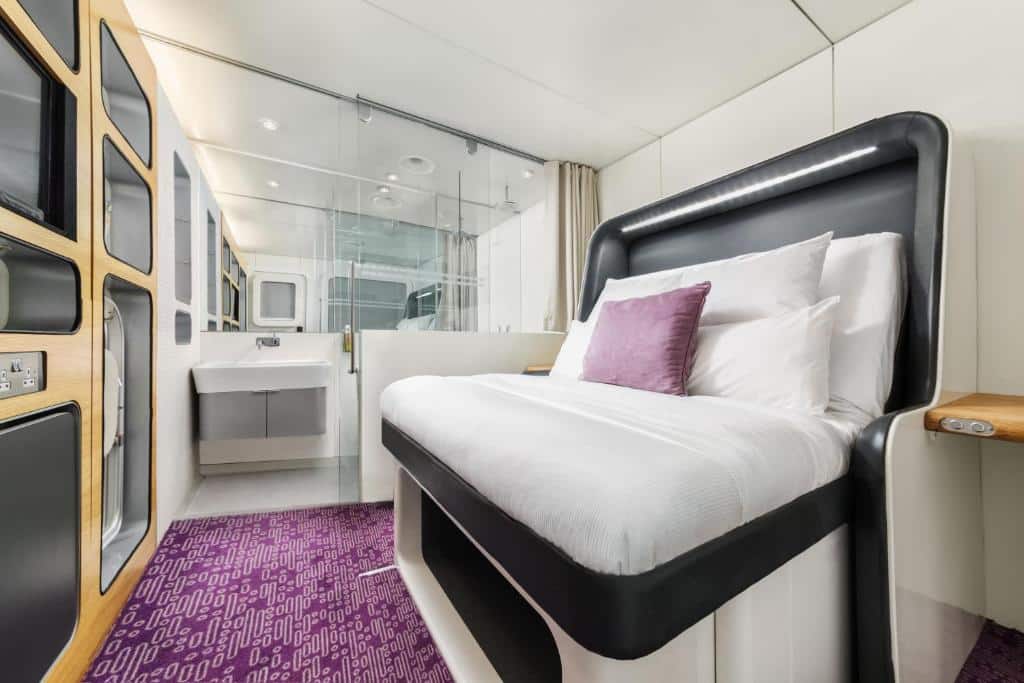 Interior de uma cabine do YOTELAIR Amsterdam Schiphol Transit Hotel, de 10 m², com uma cama alta com repartição para colocar utensílios abaixo, um balcão pequeno no lado direito e, na frente da cama, tem uma parede com TV e outras repartições. Ao lado esquerdo, há o banheiro. Imagem para ilustrar o post de hotéis perto do aeroporto de Amsterdam