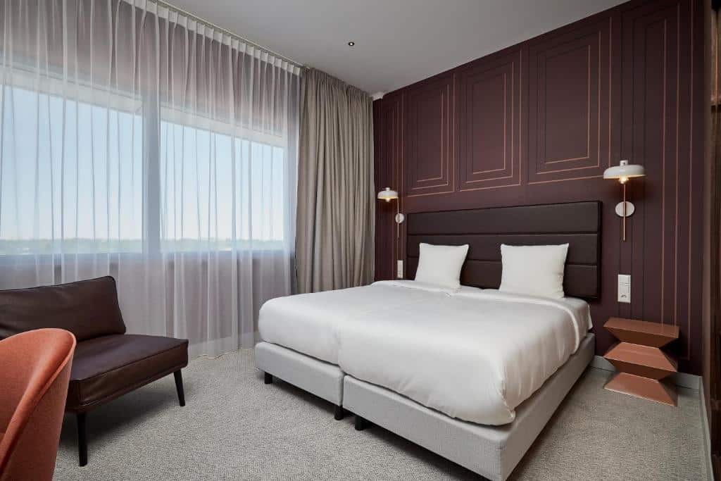 Quarto do Corendon Plaza Amsterdam Schiphol Airport Hotel, de 21 m², com uma cama de casal, mesinha ao lado, uma poltrona a frente e duas janelas ao lado com cortinas a frente. Imagem para ilustrar o post hotéis perto do aeroporto em Amsterdam
