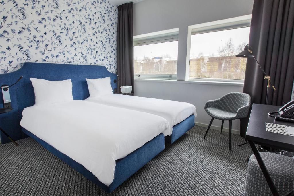 Quarto do ibis Styles Amsterdam Airport, de 24 m², com duas camas de solteiro, uma poltrona e mesa de trabalho e, no lado direito, tem duas janelas de vidro com vista para prédios históricos e árvores. Imagem para ilustrar o post de hotéis perto do aeroporto de Amsterdam