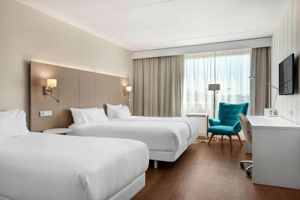 Quarto do NH Amsterdam Schiphol Airport, com duas camas, uma poltrona verde, uma mesa de trabalho, TV e uma janela ampla com vista da cidade e cortinas a frente. Imagem para ilustrar o post hotéis perto do aeroporto em Amsterdam