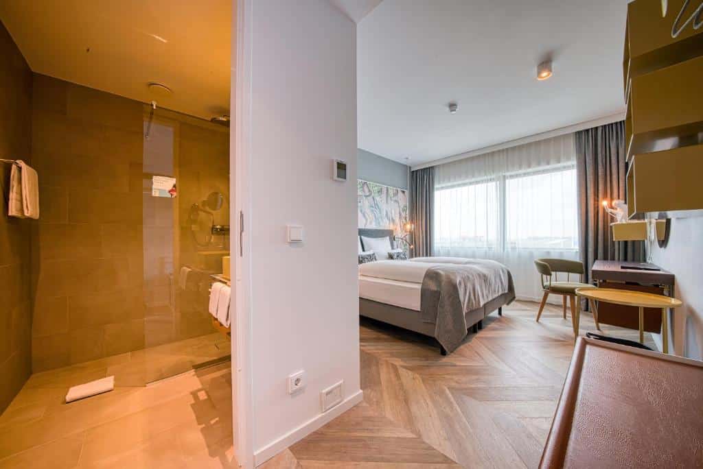 Quarto do NinetyNine Amsterdam Airport, de 19 m², com um banheiro com box de vidro e toalhas penradas na pia, e ,no lado direito, há uma cama de casal, uma mesa redonda e outra mesa de trabalho com uma cadeira