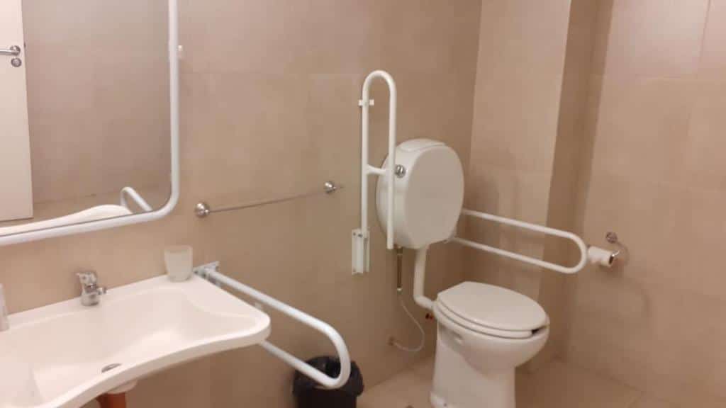 banheiro com adaptações para pessoas com deficiência como barras de apoio ao redor do vaso sanitário, cadeirinha de banho, apoio na pia com espaço embaixo no Palermo Bridge, um dos hotéis perto do aeroporto de Buenos Aires