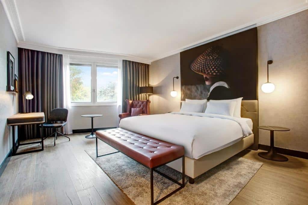 Quarto do Radisson RED London Heathrow com uma cama de casal ampla, uma janela com cortinas, o chão é de madeira, há uma poltrona e duas luminárias nas laterais da cama