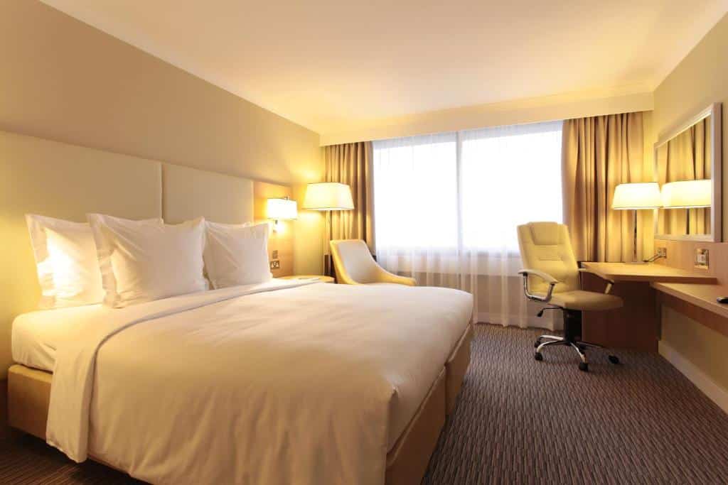 Quarto do Renaissance London Heathrow Hotel com uma cama de casal, carpete, uma janela com cortinas, uma mesa com uma cadeira giratório, há uma poltrona e um abajur de chão, para representar hotéis perto do Aeroporto de Londres Heathrow