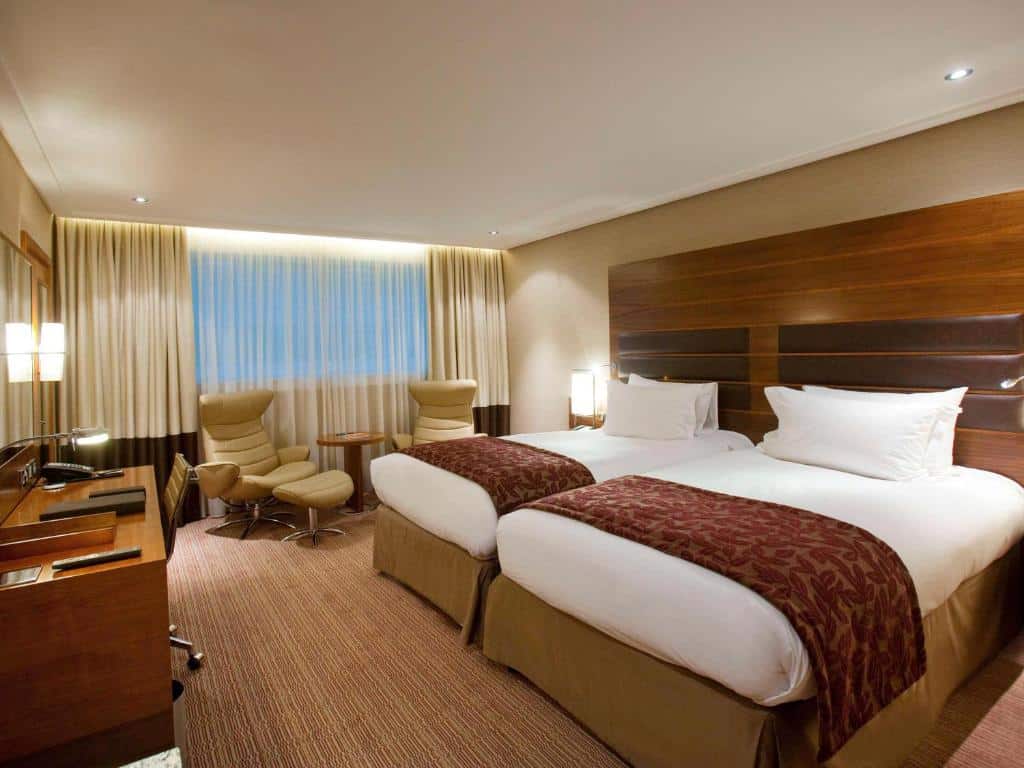 Quarto do Sofitel London Heathrow com uma cama de casal, uma de solteiro, uma janela com cortinas, carpete e uma mesa de escritório com uma cadeira de rodinha