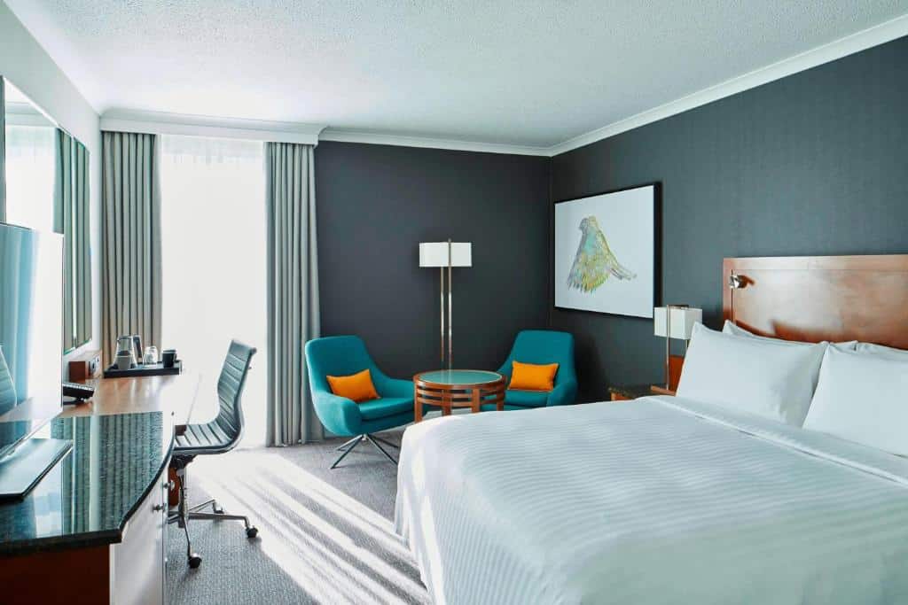 Quarto do London Heathrow Marriott Hotel com uma cama de casal, uma janela com cortinas, uma mesinha redonda com duas poltronas, chão de carpete e uma mesa de escritório